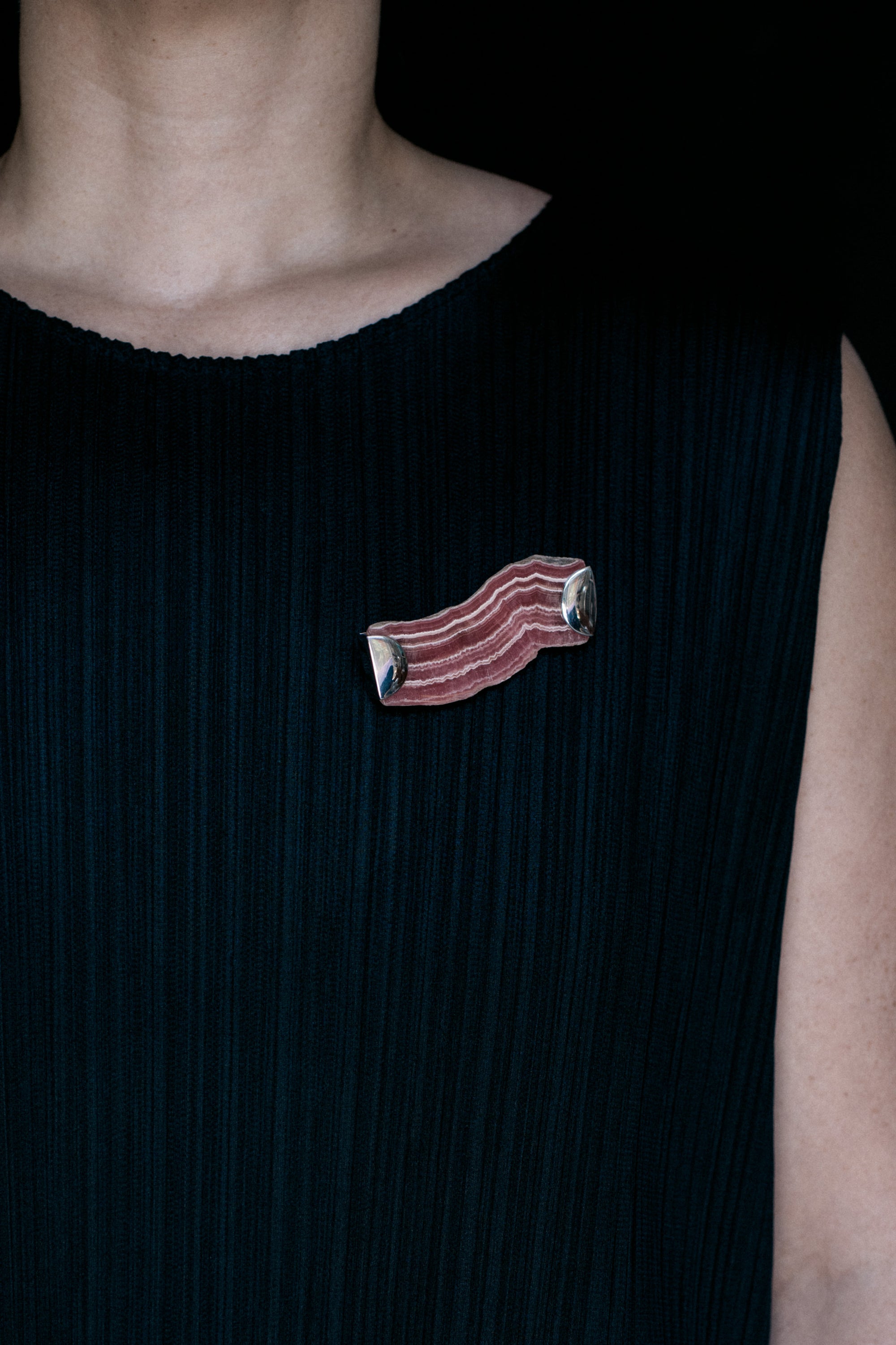 Person wearing a black sleeveless pleated dress with a bacon-shaped rhodochrosite brooch pin on a dark background