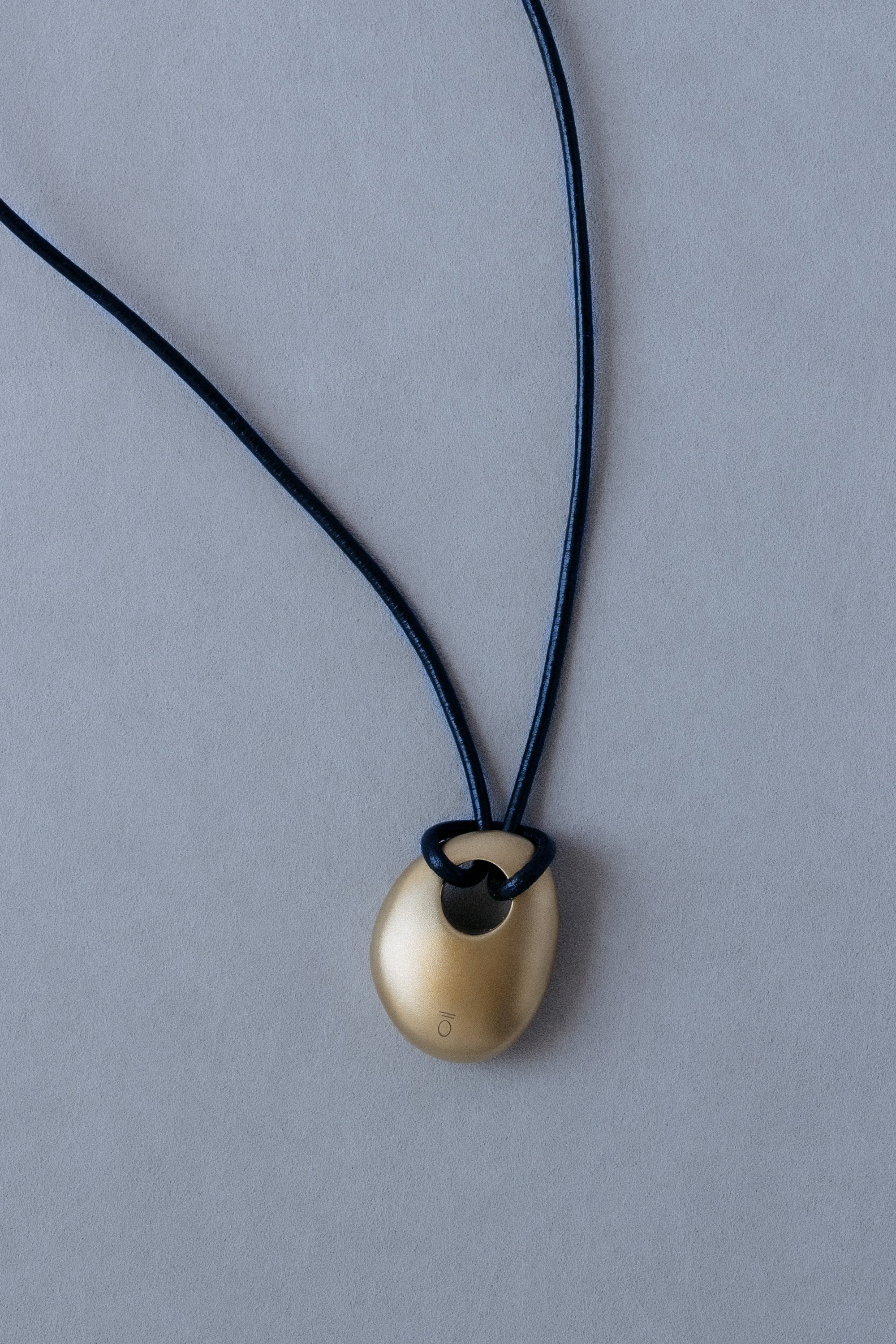 Ouvelle Brass Egg Shaped Pendant on Black Leather cord with brass tipped ends back view on grey background